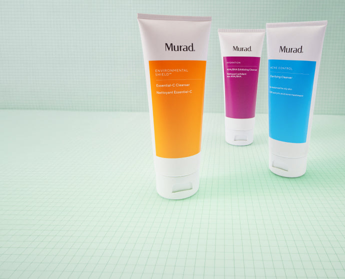 Three Murad skincare tubes standing upright on a pale mint-green gridded surface: an orange tube in front, a purple tube in the middle, and a blue tube behind. The Murad logo is visible on each tube.