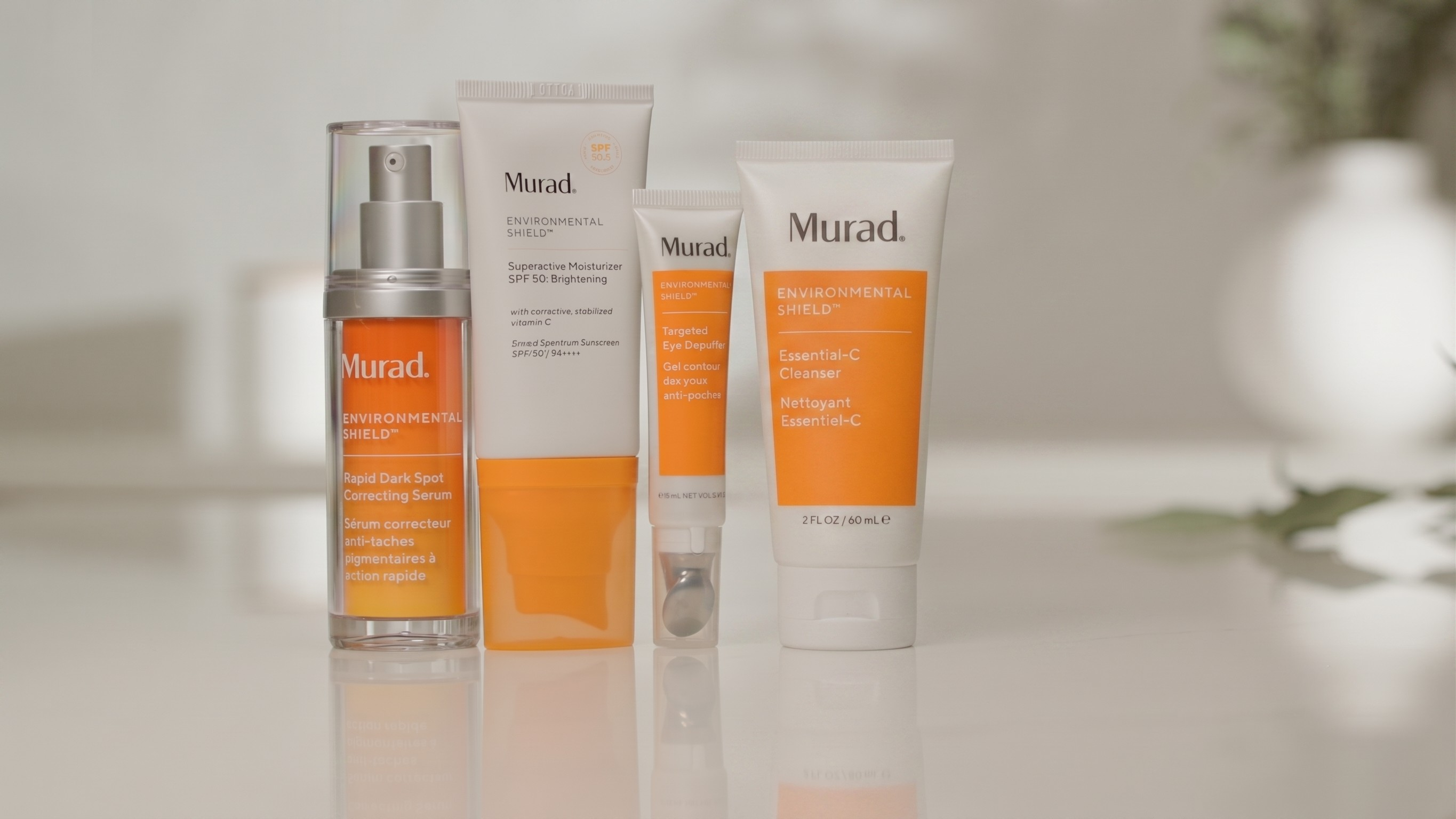 Four Murad skincare products arranged on a glossy white surface against a softly blurred light background: a clear pump bottle with orange label, a white box with orange lower section, a small white tube with orange label, and a larger white squeeze tube with orange label.